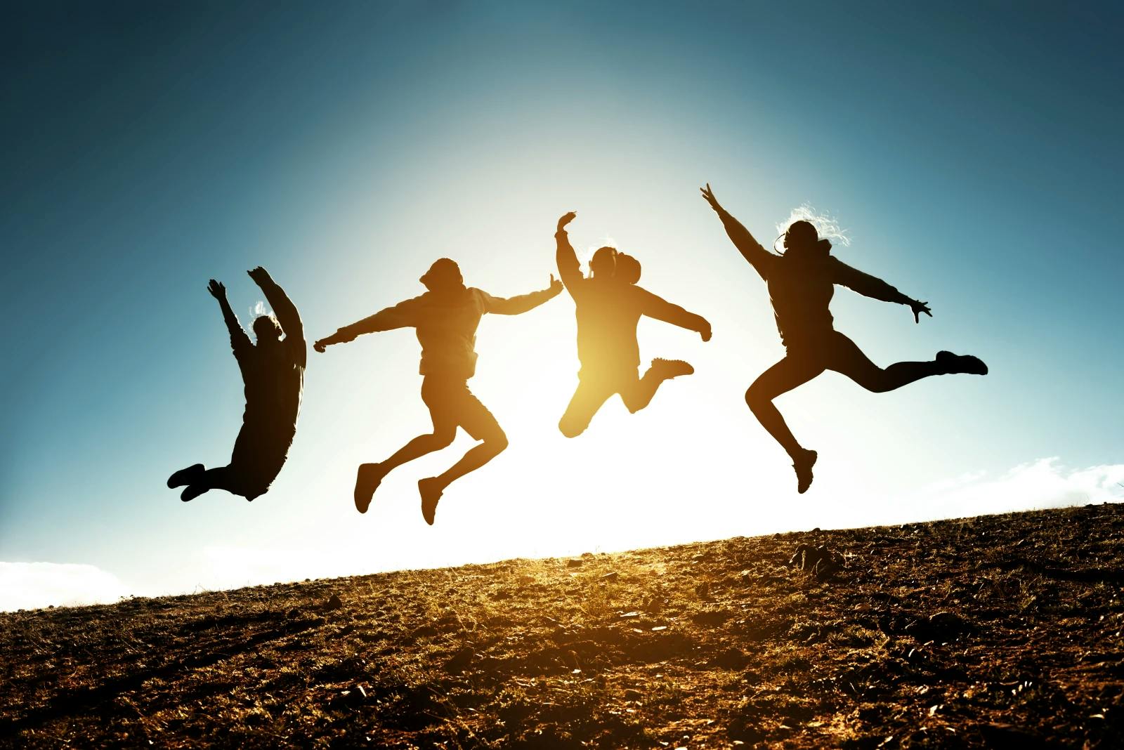 A photo of four individuals jumping for joy as silhouettes in front of a sunset on a hill.
