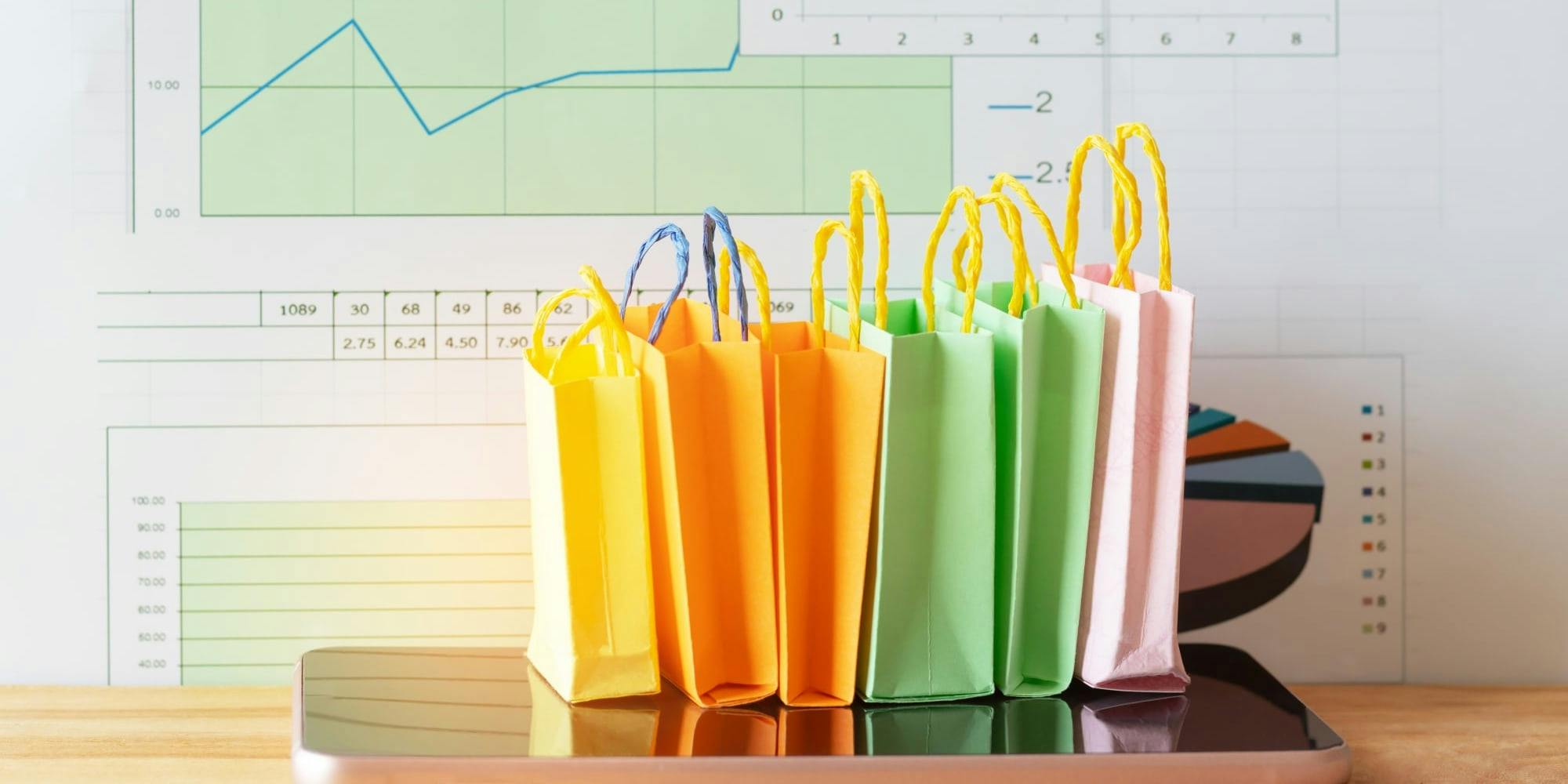 Miniature, colorful shopping bags sit on top of a glossy, pink phone. The background is of charts and graphs, somewhat like a printed Excel file.